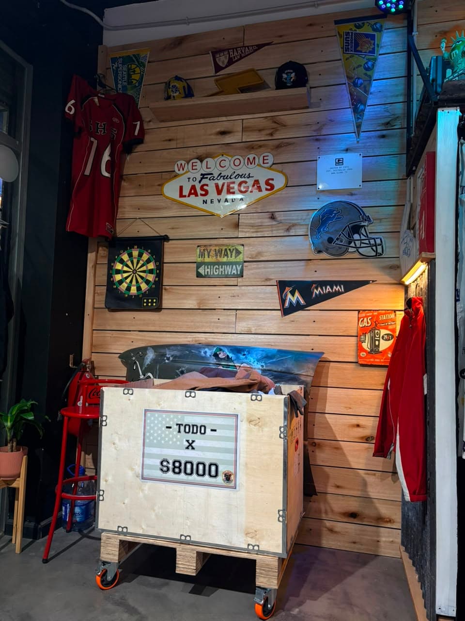 Cajón de madera con ropa suelta y el cartel Todo x $8000, bajo un cartel de Las Vegas y un casco de los Detroit Lions