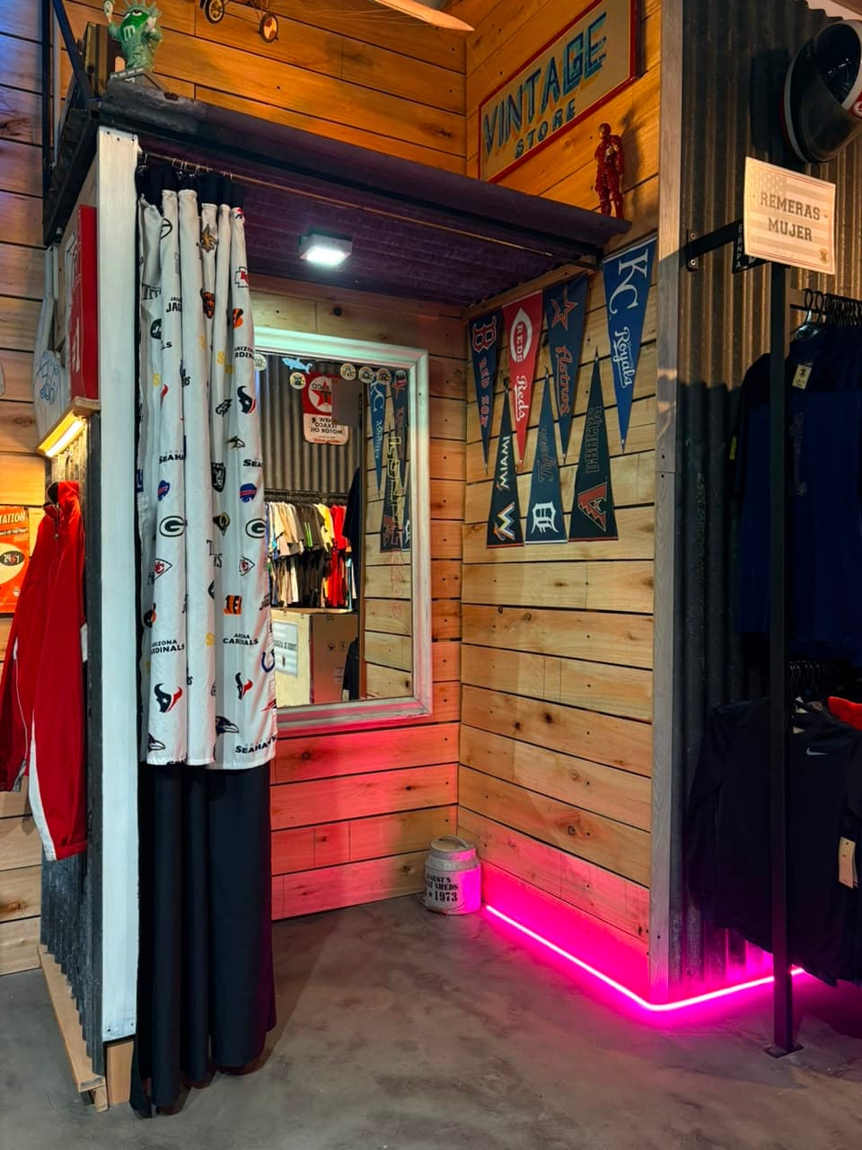 Probador del local con cortina de logos de la NFL, cartel de Vintage Store y banderines de béisbol en la pared de madera