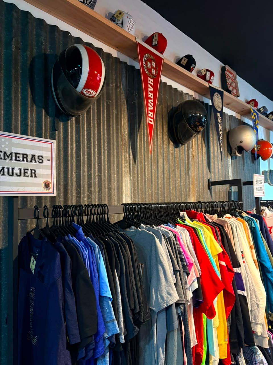 Sector de remeras de mujer con cascos vintage y un banderín de Harvard sobre la pared de chapa